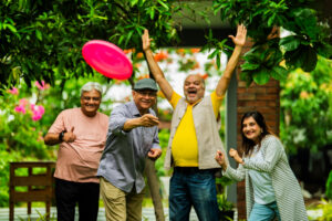 A vibrant outdoor scene showing four joyful elderly Indian friends playing frisbee. In the foreground, a man in a yellow vest and t-shirt with his arms raised in excitement watches as a man in a grey cap throws a pink frisbee towards him. To the left, another man gives a thumbs-up, and to the right, a woman smiles, ready to play. Lush green foliage and a brick structure are in the background, suggesting a garden or park setting.