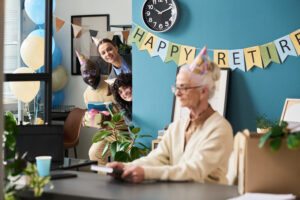 A diverse group of three younger colleagues are peeking from behind a wall, holding presents and smiling, wearing party hats, and looking towards an elderly woman in the foreground who is seated at a desk, facing away from them, appearing to be surprised. A "HAPPY RETIREMENT" banner hangs on the blue wall, along with balloons and a clock, indicating a surprise retirement party.