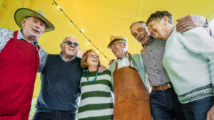 A cheerful group of six diverse senior citizens are gathered closely together, smiling and laughing under a bright yellow canopy with string lights. Two men on the left are wearing aprons, one red and one brown, and hats. The group is looking at each other or smiling towards the camera, with some putting arms around shoulders, conveying a sense of camaraderie and enjoyment at an outdoor gathering.