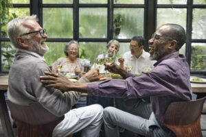 Two laughing senior men, one white with a beard and grey hair, and one Black with a short beard, are clinking wine glasses in a toast while seated at a wooden table. The white man has his hand on the other man's shoulder. In the blurred background, three other diverse senior individuals are also seated at the table, smiling and engaged in conversation. The setting appears to be a restaurant or café with large windows looking out onto greenery.