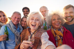 A group of diverse, smiling people of various ages, including several seniors and younger adults, are crowded together taking a selfie outdoors under bright sunlight. The woman in the center foreground is laughing with her mouth open, holding a glass of white wine, and her hand is extended as if holding the camera. Others are also smiling and some hold drinks, conveying a joyful and celebratory gathering.