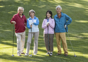 Four smiling senior golfers, two couples, stand side-by-side on a green golf course, each holding a golf club. From left to right: a white man in a red polo, a white woman in a blue and white jacket, an Asian woman in a pink jacket, and an Asian man in a blue jacket. All are looking at the camera, appearing happy and relaxed.