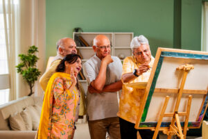A group of four diverse senior adults are gathered around an easel, looking intently at a painting on it. One elderly man in a yellow patterned shirt is pointing at the canvas with a brush, appearing to instruct or demonstrate. Another man in a light grey shirt has his hand on his chin, looking thoughtful. An elderly woman in a colorful patterned outfit and yellow shawl has her hand raised in a gesture of surprise or admiration. A fourth man in a yellow shirt stands behind them. The setting appears to be a brightly lit indoor space, possibly a living room or art studio, with a couch and shelves in the background.