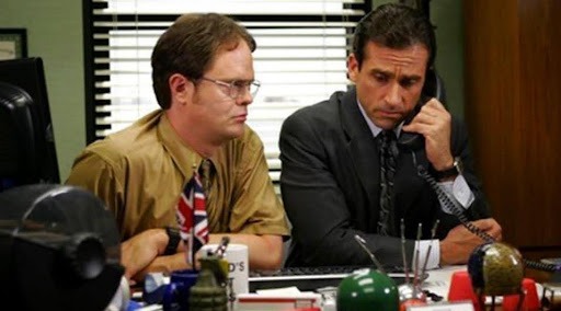 Two men are seated at an office desk. The man on the left, wearing a mustard-colored shirt and glasses, looks intently to his left. The man on the right, in a dark suit and tie, holds a black telephone receiver to his ear with a serious expression, looking down at the desk. The desk is cluttered with various office items, including a mug, a small flag, and decorative objects.