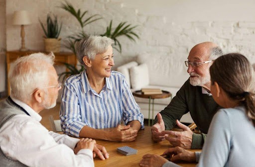 Four diverse senior adults are seated around a wooden table, engaged in lively conversation, possibly playing cards as a deck of cards is on the table. A woman in a striped shirt is smiling and looking at a bearded man in glasses and a green sweater who is gesturing with his hands as he speaks. Another man with white hair and glasses is listening intently on the left, and a fourth person is partially visible on the right. The setting appears to be a comfortable indoor space with plants and furniture in the blurred background.