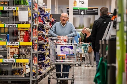 An elderly man wearing a light blue sweater and glasses is pushing a full shopping cart down an aisle in a grocery store, looking towards the camera with a neutral expression. The aisle is lined with shelves of various products, some marked with "1/2 Price" signs. Other shoppers are visible in the blurred background.