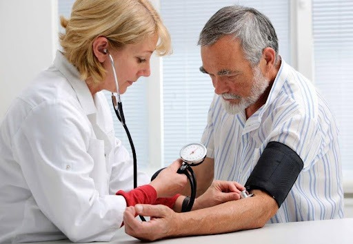 A medical professional, a woman with blonde hair wearing a white lab coat and a stethoscope around her neck, is taking the blood pressure of a senior man with a beard. She is looking intently at the gauge while adjusting the cuff on his arm. The man is looking down at his arm, and they are both seated indoors in what appears to be a clinical setting.