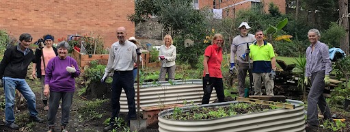 A diverse group of about nine people, including several seniors and some younger individuals, are gathered in a community garden, posing and smiling at the camera. They are dressed in casual clothes and some wear gloves, standing near raised garden beds, with a brick building and lush greenery in the background. The scene suggests a collaborative gardening or community activity.