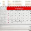 A close-up view of a desk with a white coffee cup on the left and a white desk calendar with red binding and header in the center. The calendar shows a month starting on a Tuesday and ending on the 31st, with some dates highlighted in red. To the right of the calendar, there's a holder with colored pencils. The background is a blurred office environment with windows.