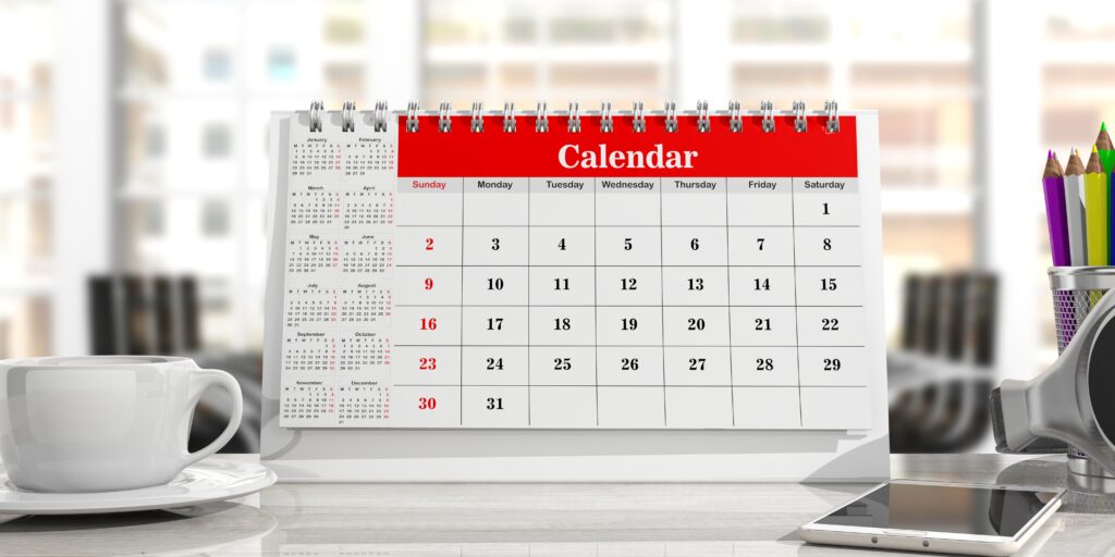 A close-up view of a desk with a white coffee cup on the left and a white desk calendar with red binding and header in the center. The calendar shows a month starting on a Tuesday and ending on the 31st, with some dates highlighted in red. To the right of the calendar, there's a holder with colored pencils. The background is a blurred office environment with windows.