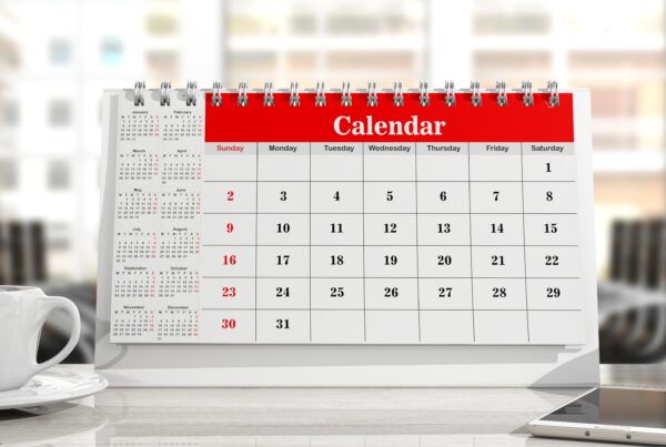 A close-up view of a desk with a white coffee cup on the left and a white desk calendar with red binding and header in the center. The calendar shows a month starting on a Tuesday and ending on the 31st, with some dates highlighted in red. To the right of the calendar, there's a holder with colored pencils. The background is a blurred office environment with windows.