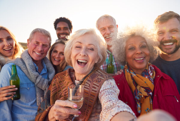 A group of diverse, smiling people of various ages, including several seniors and younger adults, are crowded together taking a selfie outdoors under bright sunlight. The woman in the center foreground is laughing with her mouth open, holding a glass of white wine, and her hand is extended as if holding the camera. Others are also smiling and some hold drinks, conveying a joyful and celebratory gathering.