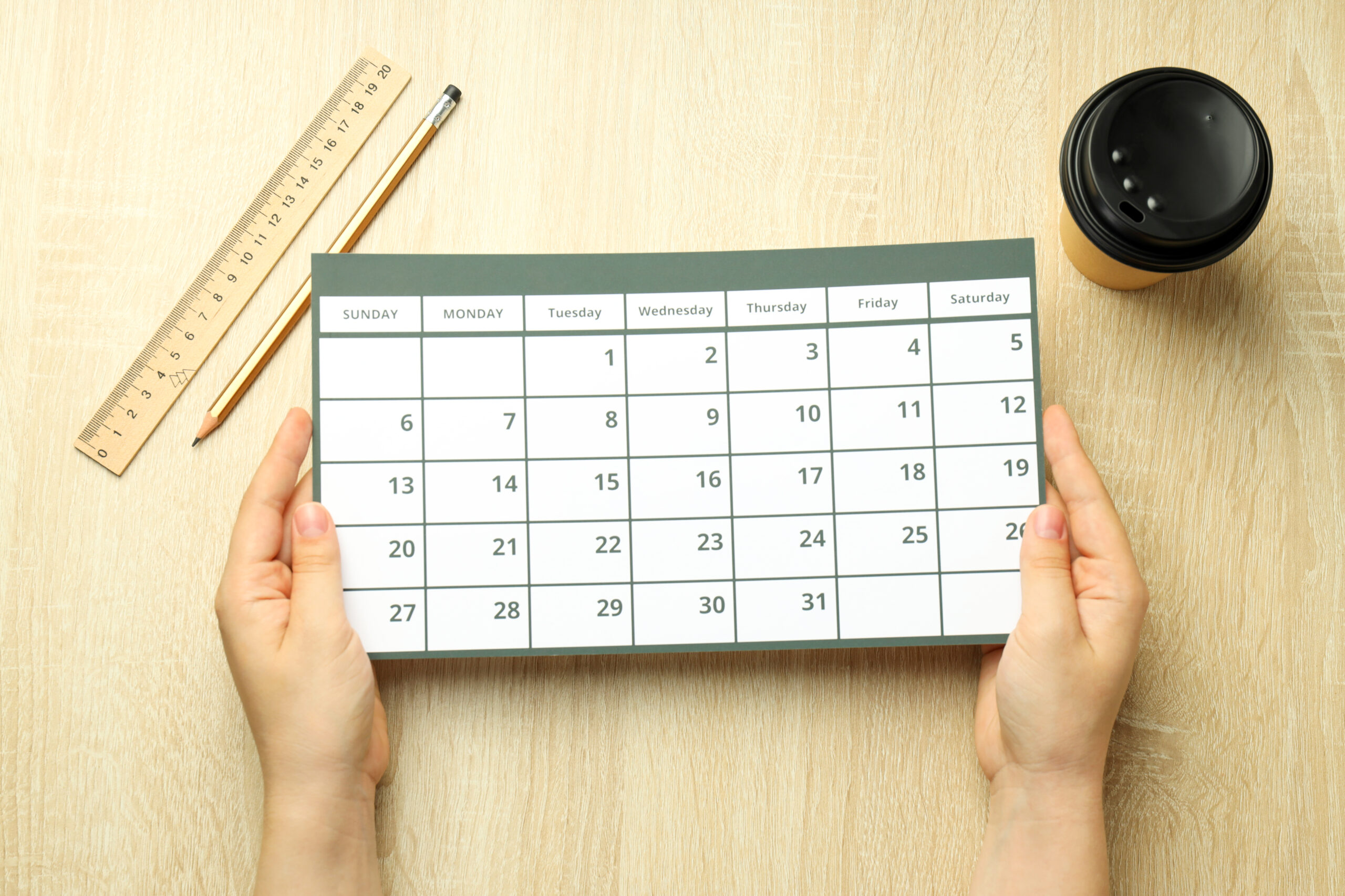 A pair of hands holds up a white monthly calendar page, likely for a month with 31 days starting on a Tuesday. The calendar is centered on a light wooden surface, with a ruler and pencil to its left and a black disposable coffee cup to its upper right.