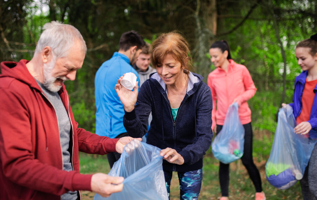 A diverse group of people, including two smiling seniors in the foreground, are actively picking up litter in a wooded area. An elderly man with a white beard, wearing a red hoodie, holds open a clear blue trash bag. Beside him, an elderly woman with reddish-brown hair, wearing a dark blue hoodie and patterned leggings, smiles as she drops a piece of white litter into the bag. Several younger individuals are visible in the background, also holding trash bags, suggesting a group environmental cleanup effort.