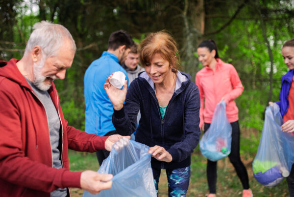 A diverse group of people, including two smiling seniors in the foreground, are actively picking up litter in a wooded area. An elderly man with a white beard, wearing a red hoodie, holds open a clear blue trash bag. Beside him, an elderly woman with reddish-brown hair, wearing a dark blue hoodie and patterned leggings, smiles as she drops a piece of white litter into the bag. Several younger individuals are visible in the background, also holding trash bags, suggesting a group environmental cleanup effort.