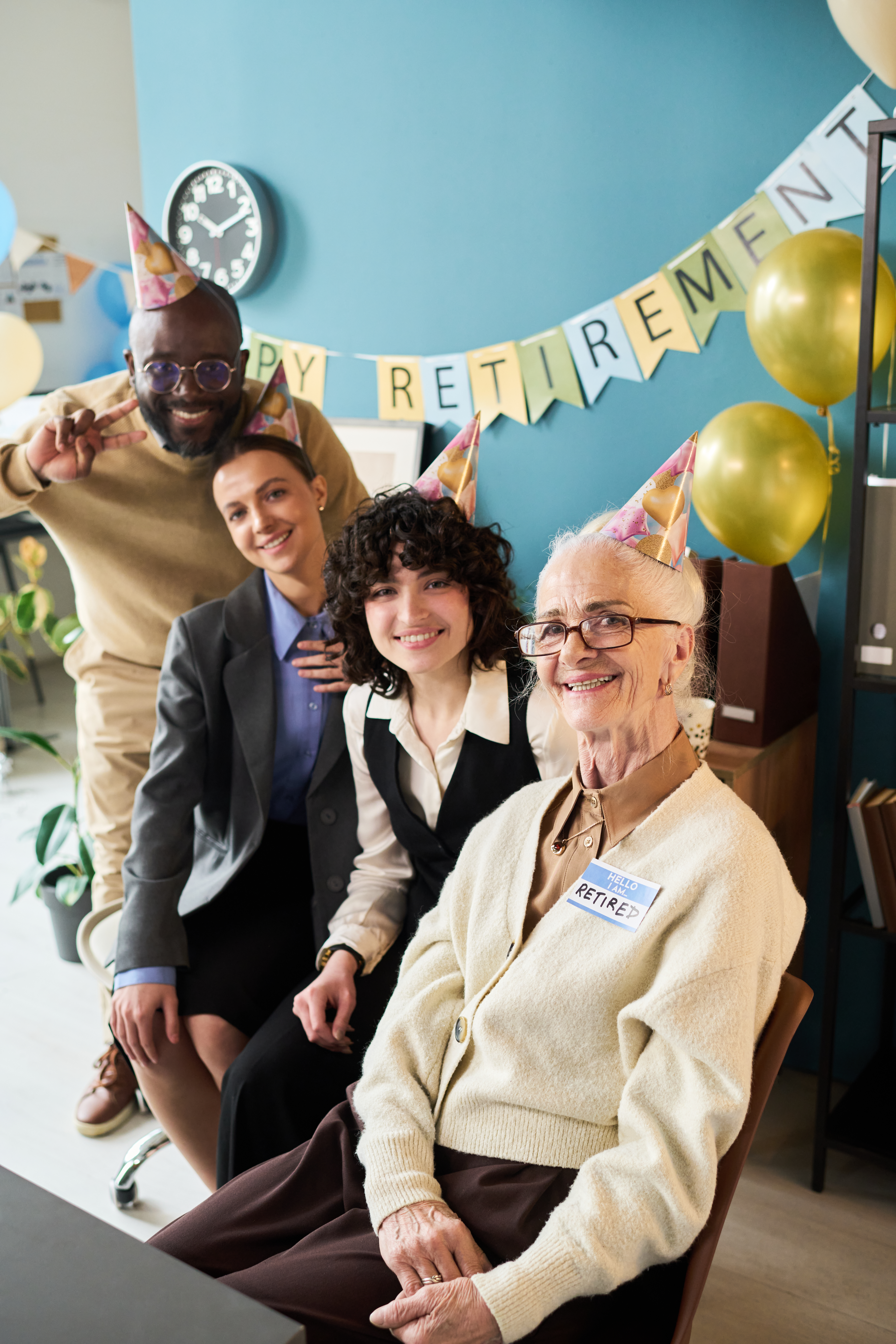 A joyous group of four colleagues, including a senior Caucasian woman, are celebrating a retirement. The senior woman, wearing a light-colored cardigan and a "RETIRED" nametag, is seated in the foreground, smiling broadly and wearing a party hat. Behind her, three younger, diverse colleagues also wear party hats and smile at the camera, with one man flashing a peace sign. Balloons and a "HAPPY RETIREMENT" banner decorate the light blue office background, indicating a festive atmosphere.