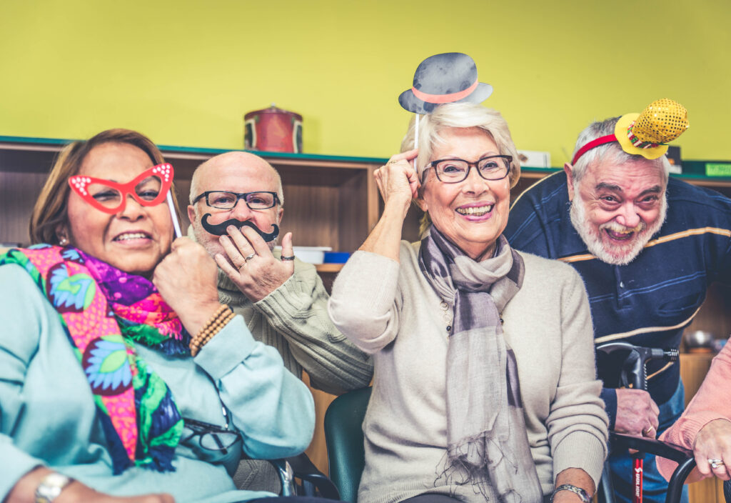 A diverse group of four joyful senior citizens is gathered together, wearing fun novelty props. From left to right: an elderly woman with red cat-eye glasses, a bald man holding a fake mustache, a smiling woman with glasses and a scarf holding a tiny top hat on a stick above her head, and an elderly man with a beard wearing a yellow novelty hat. They are all laughing and looking at the camera, suggesting a lively and fun social event.
