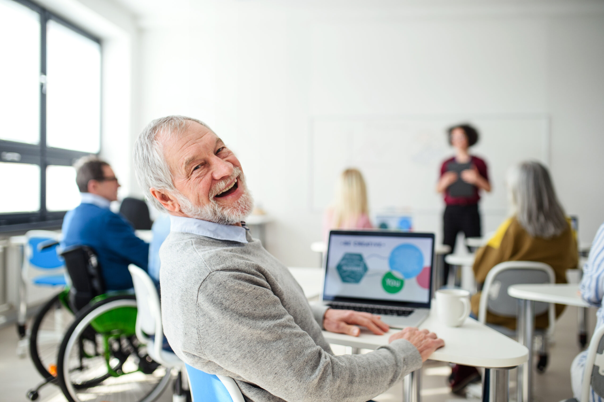 A happy, smiling senior man with a beard and grey hair looks back at the camera from his desk in a classroom setting. In front of him is an open laptop displaying colorful graphics. In the background, other adults, including some in wheelchairs, are seated at desks, and a woman appears to be leading a presentation at a whiteboard. The room is bright with natural light from large windows.