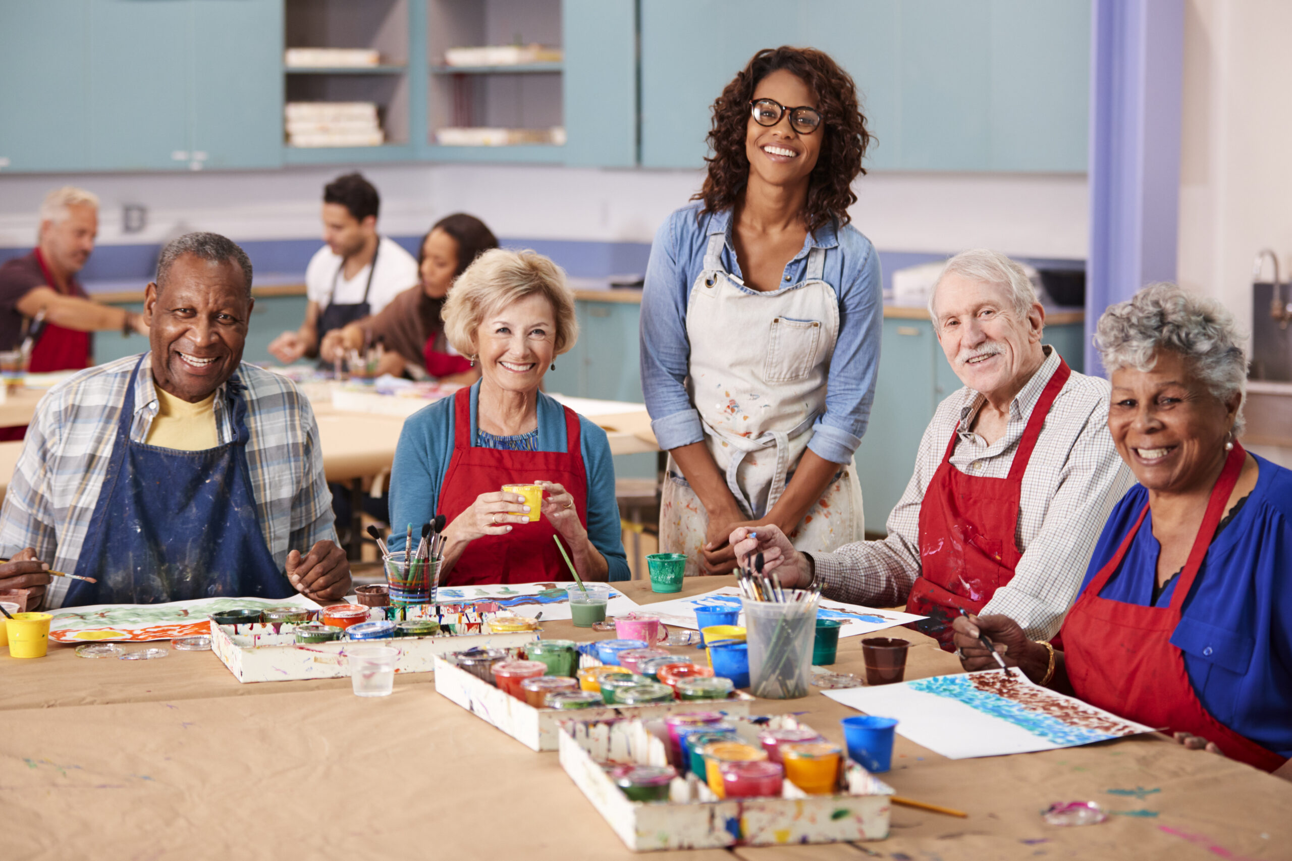 A diverse group of five smiling senior citizens, wearing colorful aprons, are seated at a long table covered in brown paper, engaged in an art class. They are painting with various paints and brushes. A smiling African American woman, likely the instructor, stands behind them, wearing an apron and glasses. Other people are visible in the blurred background of the bright classroom.
