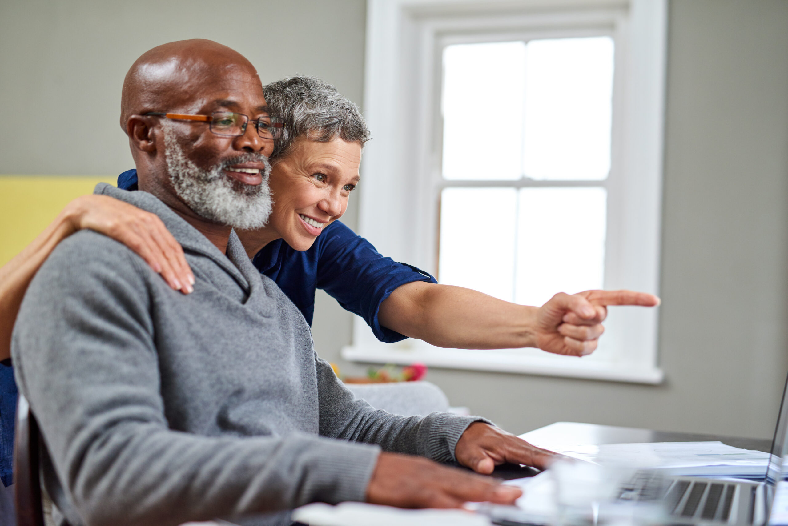 A diverse senior couple is looking at a laptop together. A smiling Black man with a white beard and glasses is seated at the computer, while a smiling white woman with short grey hair leans over his shoulder, pointing at the screen. Her arm is around his shoulder, suggesting a close and cooperative interaction. A window is visible in the background.