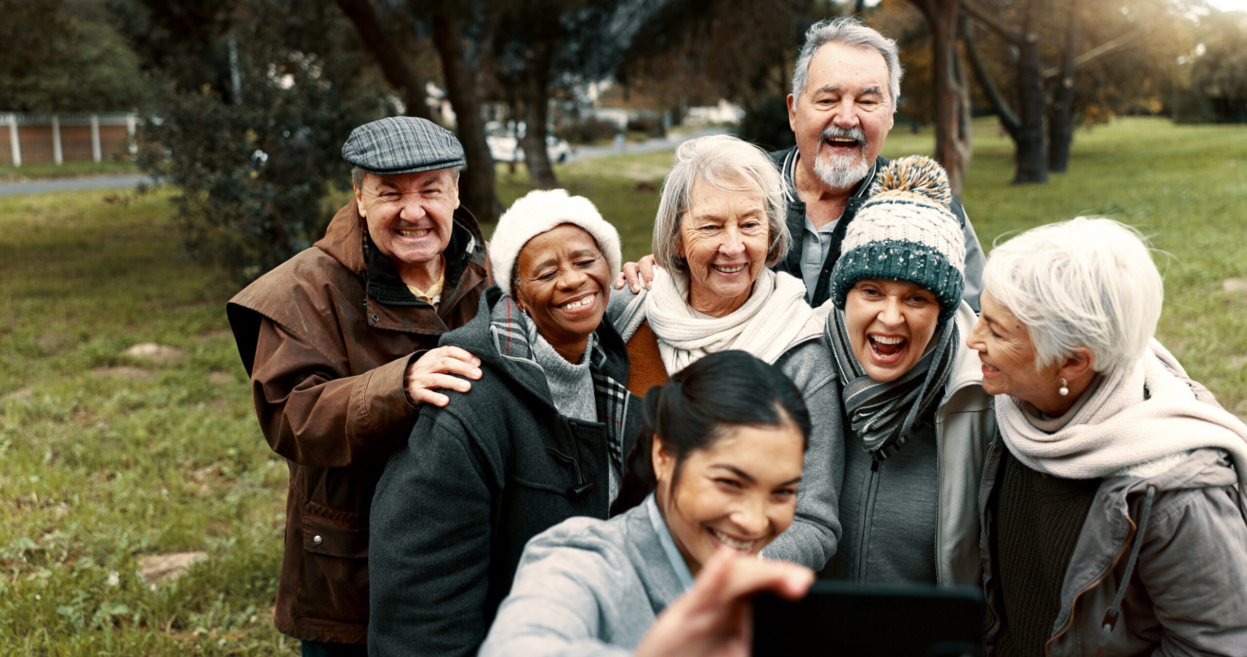 A cheerful group of six diverse senior citizens are posing closely together for a selfie, being taken by a smiling younger woman in the foreground whose arm and phone are visible. The seniors are laughing and smiling, wearing warm clothing like hats, scarves, and jackets, suggesting an outdoor setting in cooler weather. Trees and a grassy park area are blurred in the background.