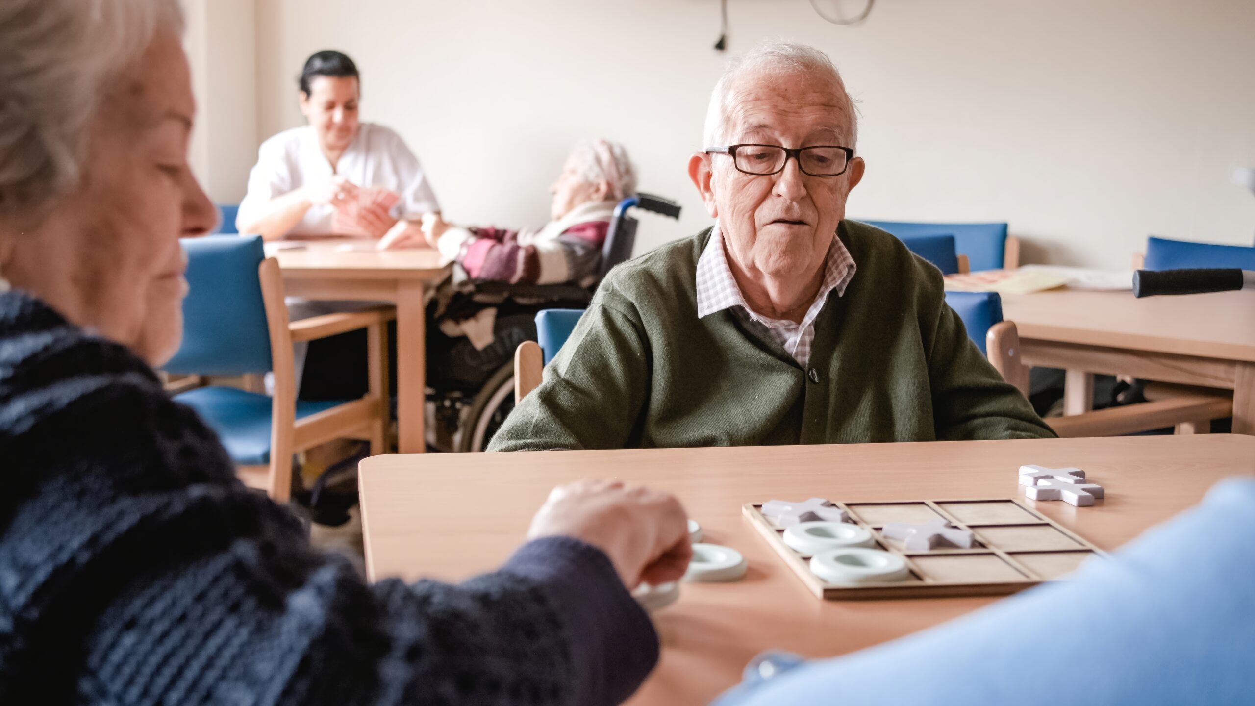 An elderly man wearing glasses and a green cardigan sits at a wooden table, looking intently at a board game in front of him. Opposite him, another elderly person, partially visible, has their hand on a game piece. In the background, other elderly individuals and a caregiver are present in a room that appears to be a common area in a care facility.