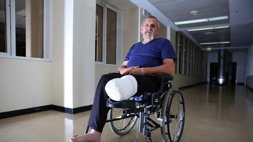 A middle-aged man with a beard, wearing a blue t-shirt and dark pants, sits in a wheelchair in a hospital hallway. His right leg is bandaged and elevated, indicating an injury or recent surgery. He is looking directly at the camera with a somber expression. The hallway is empty, with plain walls and overhead lighting.