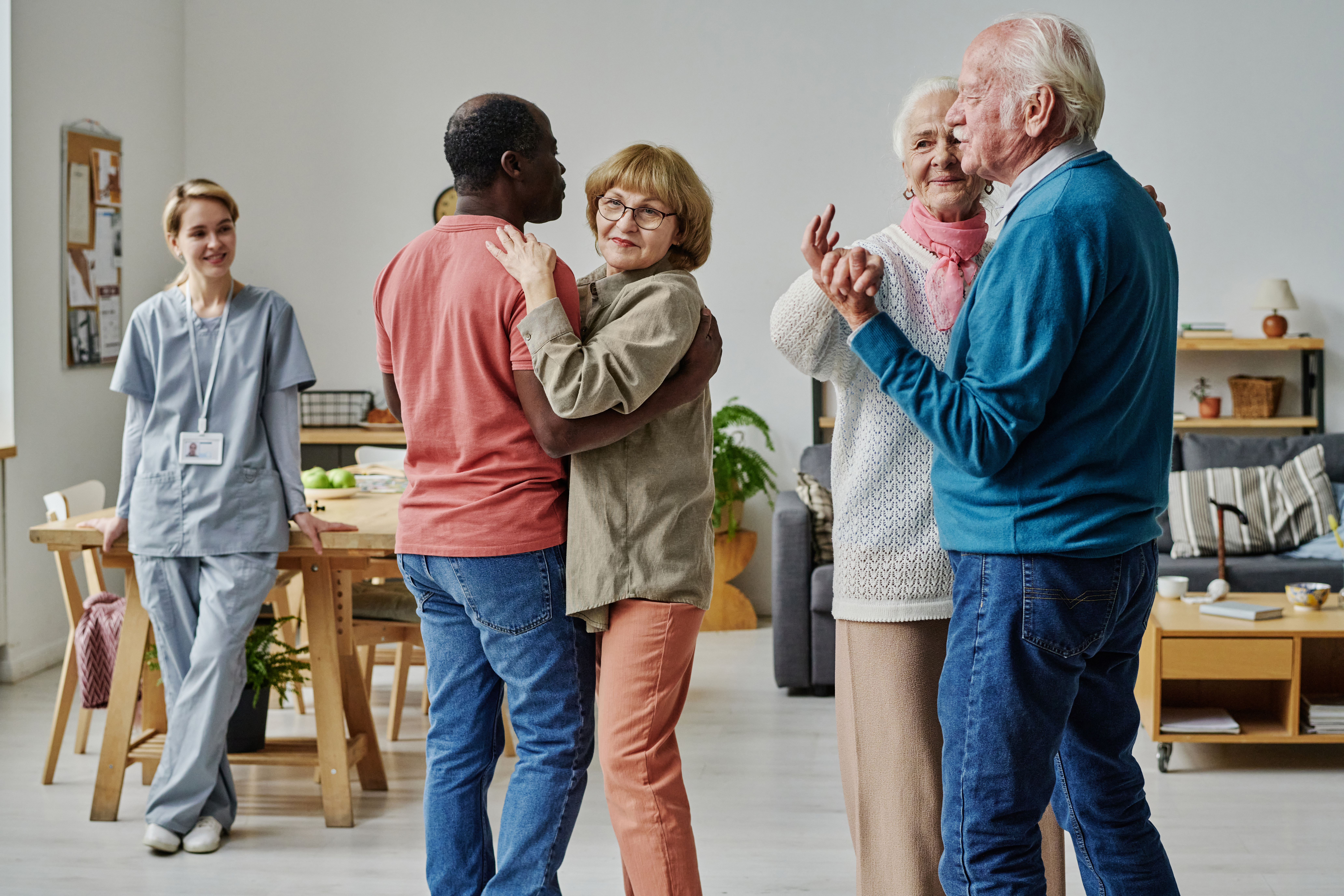 Two elderly couples are dancing in a brightly lit room, with one couple embracing in the foreground and the other holding hands and smiling. In the background, a female caregiver in blue scrubs stands by a wooden table, observing the scene with a smile. The room features modern furniture, including a sofa and shelves, suggesting a comfortable communal living space.