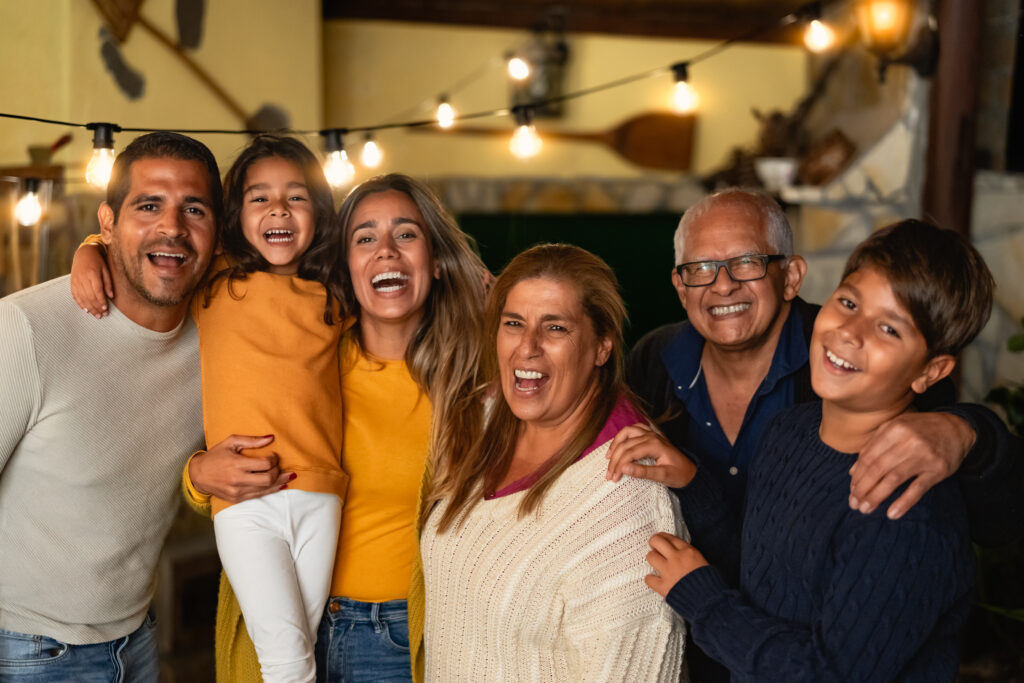 A joyous multi-generational family of six, including children, adults, and seniors, smiles and laughs at the camera. They stand close together, with festive string lights visible in the background.
