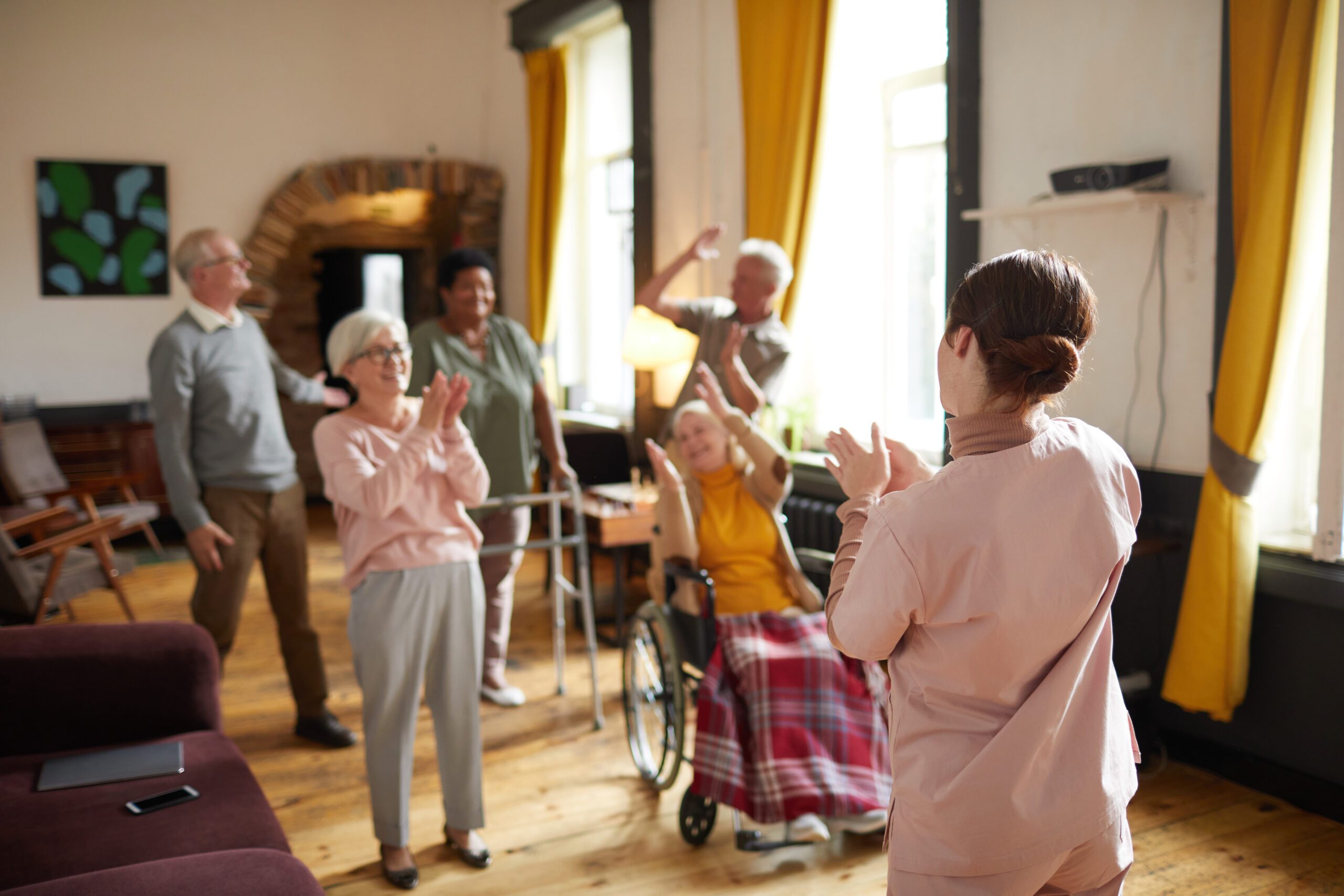 A diverse group of five elderly people, some standing and one in a wheelchair, are enthusiastically clapping and raising their hands in a brightly lit room with yellow curtains. They are facing away from the camera, towards a caregiver in light pink scrubs who is also clapping, seemingly leading an activity or exercise session.