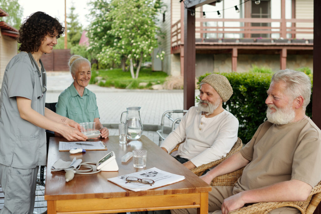 A smiling young nurse in light grey scrubs is serving food or medication to a group of three happy senior citizens seated around a wooden outdoor table. One elderly woman in a green shirt smiles at the nurse, while two bearded elderly men, one wearing a beanie, look contentedly at the nurse or the table. The setting is an outdoor patio with a building and greenery in the background.