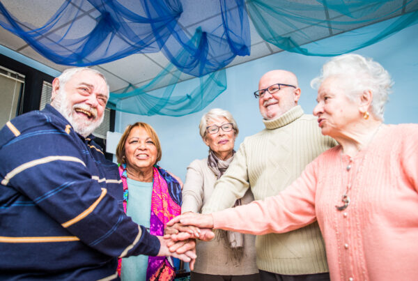 Five diverse senior citizens are gathered in a huddle, with their hands stacked in the center, all smiling and appearing joyful. They are in a room with light blue walls and decorative blue netting hanging from the ceiling.