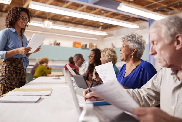 A diverse group of senior adults are attending a class, likely an IT or computer literacy course, in a brightly lit room. A smiling African American woman with curly hair and glasses, presumably the instructor, stands at the front, holding papers and looking at the class. Several seniors are seated at tables with laptops, some are looking at papers, and one elderly man in the foreground is writing. The atmosphere appears engaged and educational.